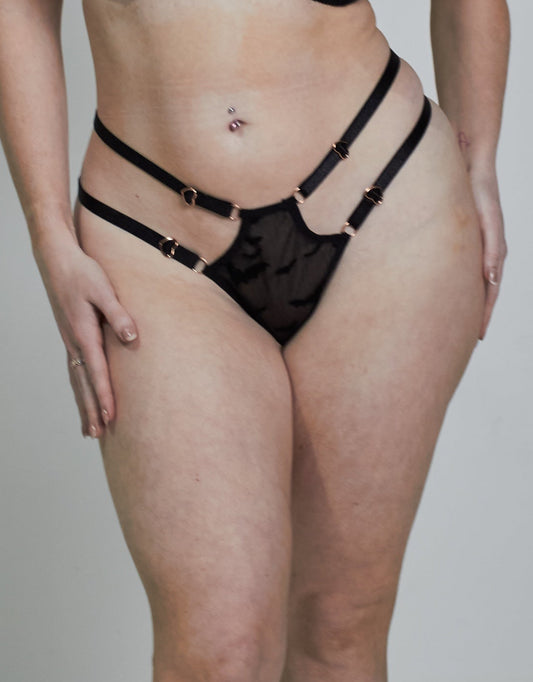A woman modeling a black lace lingerie set consisting of a bra and matching garter belt and panties, standing against a plain light gray studio background.