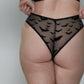 A woman stands with her back to the camera wearing a black lingerie set consisting of a black bra and black sheer mesh panties with a bat print, paired with white socks, against a plain light gray wall.