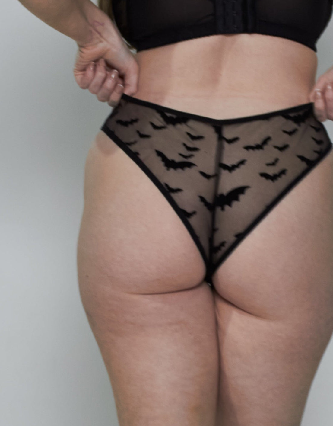 A woman stands with her back to the camera wearing a black lingerie set consisting of a black bra and black sheer mesh panties with a bat print, paired with white socks, against a plain light gray wall.
