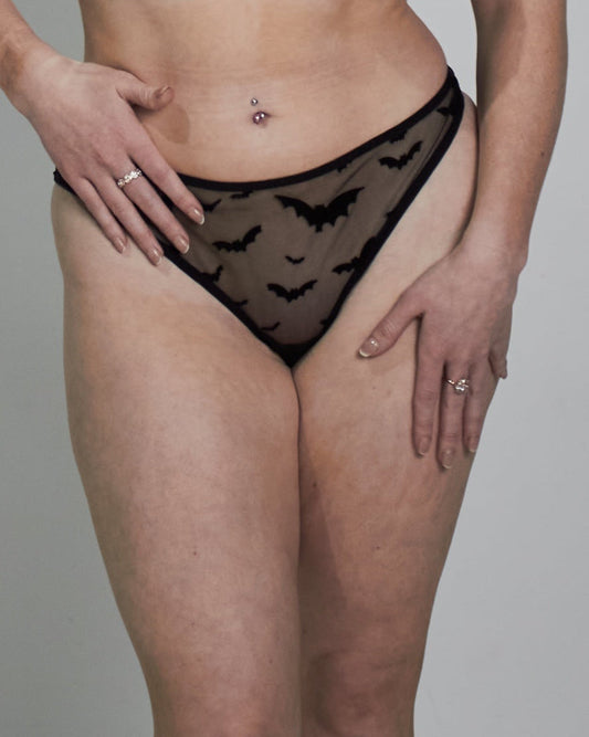 A woman modeling a black sheer lace lingerie set consisting of a bra and matching panties, standing against a plain light gray studio background.