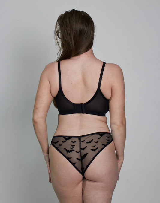 A woman stands with her back to the camera wearing a black lace bra and matching lace underwear against a plain light gray studio background.