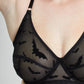 A woman wearing a black sheer lace lingerie bra with bat motifs and matching bottoms, photographed against a plain light gray studio background.