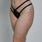 A side-profile studio photo of a woman modeling black sheer lace lingerie, including a bra and thong-style underwear, with white socks, against a plain light gray wall and floor.