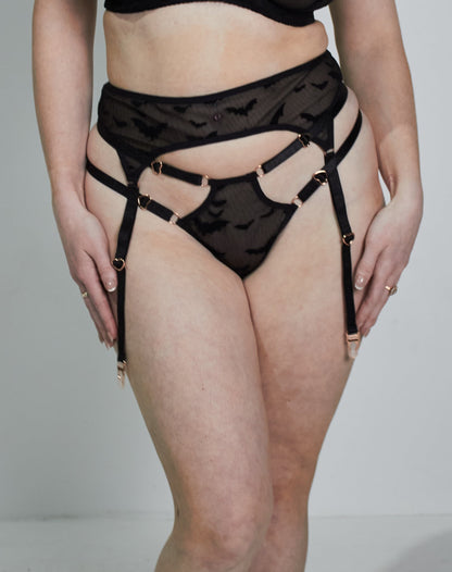 A woman modeling a black lace lingerie set consisting of a bra, garter belt, and panties with attached garters, photographed against a plain light gray studio background.