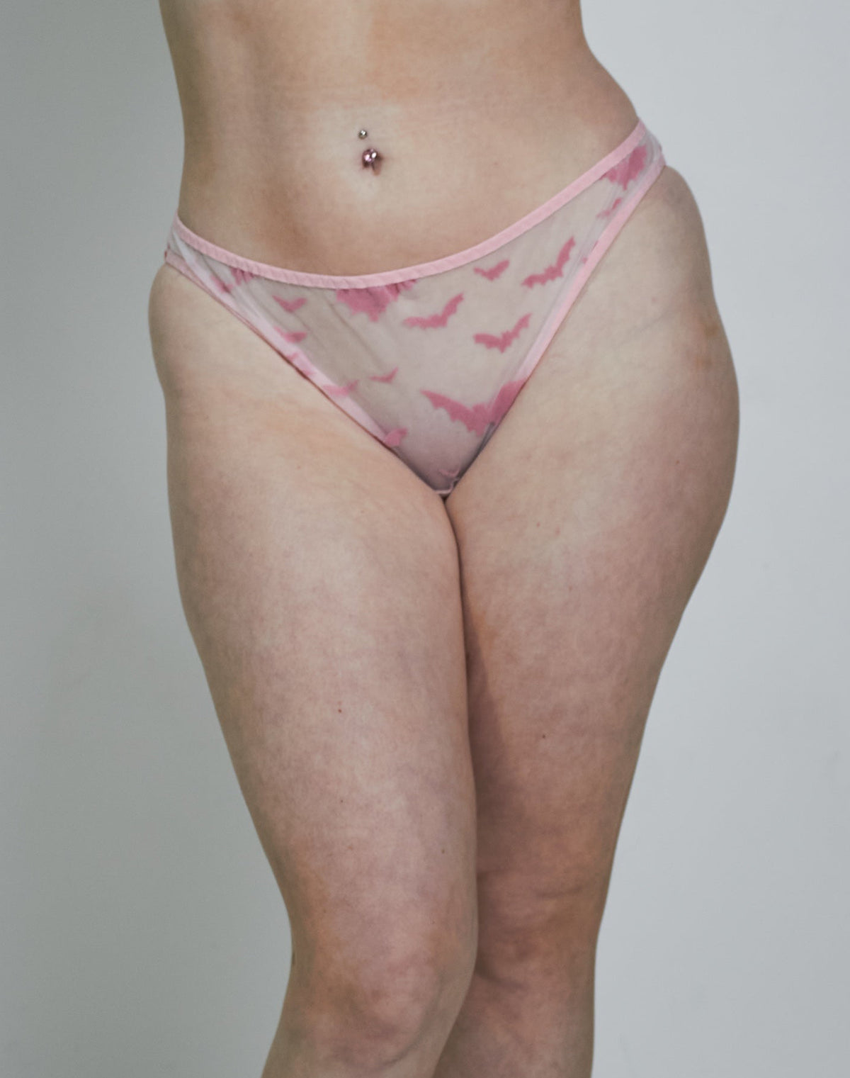A woman modeling a light pink sheer mesh lingerie set with a darker pink abstract pattern, consisting of a triangle bra and matching thong, paired with white socks against a plain light gray studio background.