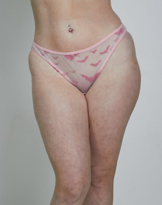 A woman modeling a light pink sheer mesh lingerie set with a darker pink abstract pattern, consisting of a triangle bra and matching thong, paired with white socks against a plain light gray studio background.