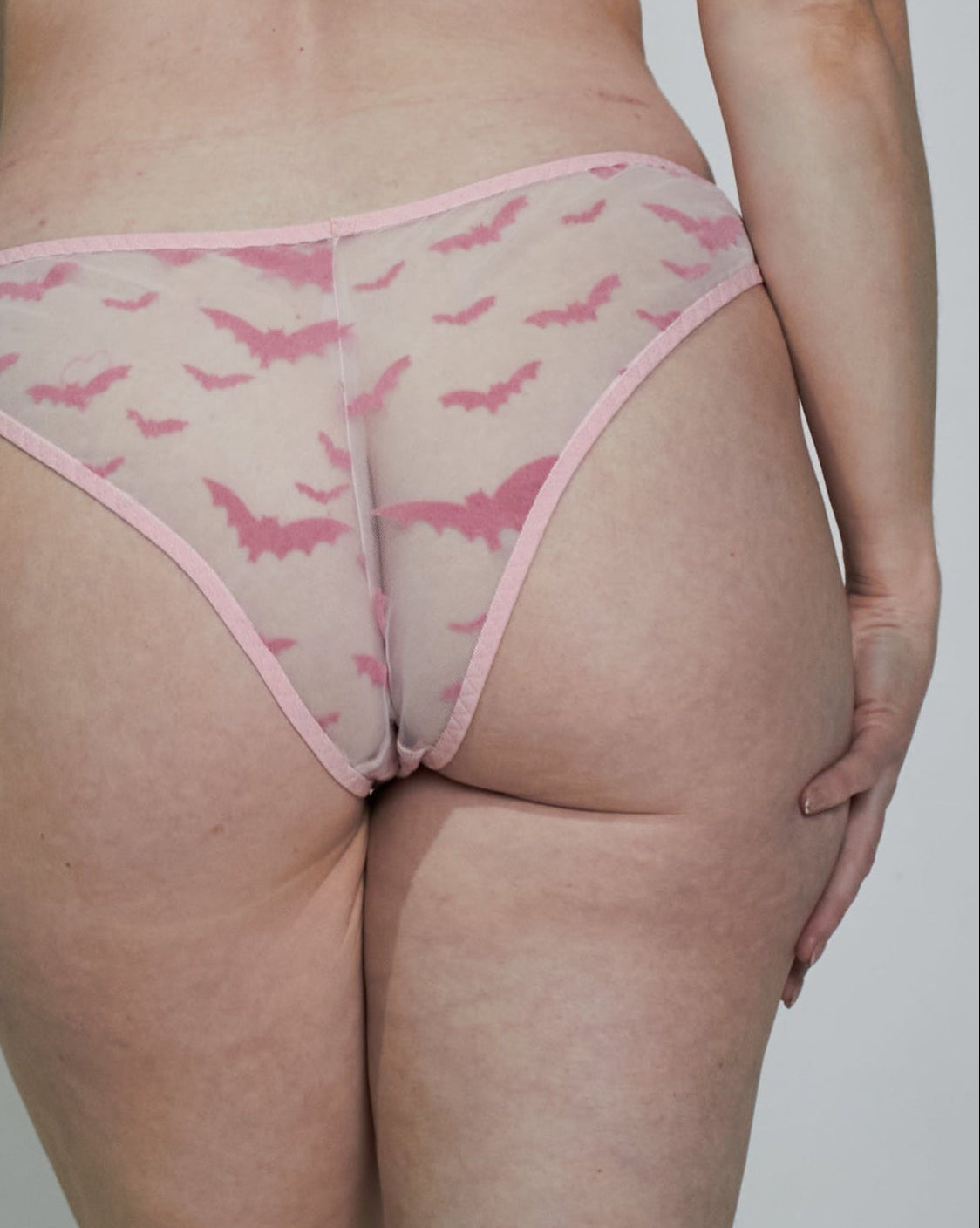 A woman shown from the back wearing a light pink lingerie set with a subtle pattern, standing against a plain light gray studio background.