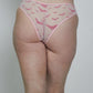 A woman shown from behind wearing a light pink lingerie set with sheer mesh and a small pink bird pattern on the thong, against a plain light gray studio background.