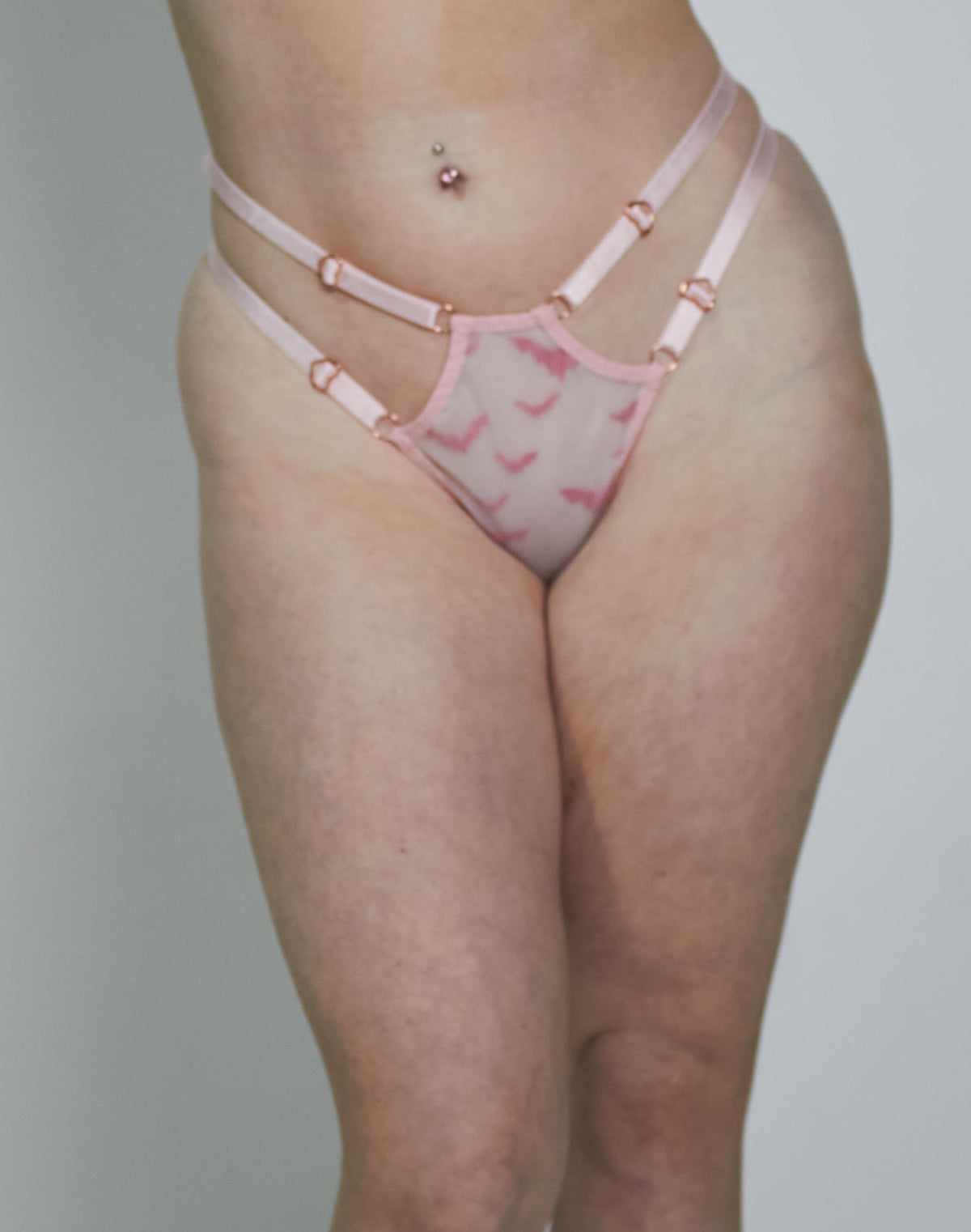 A woman modeling a light pink lingerie set with a darker pink abstract pattern, consisting of a bra and matching thong, set against a plain light gray studio background.