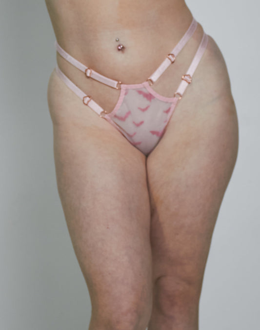 A woman modeling a light pink lingerie set with a darker pink abstract pattern, consisting of a bra and matching thong, set against a plain light gray studio background.