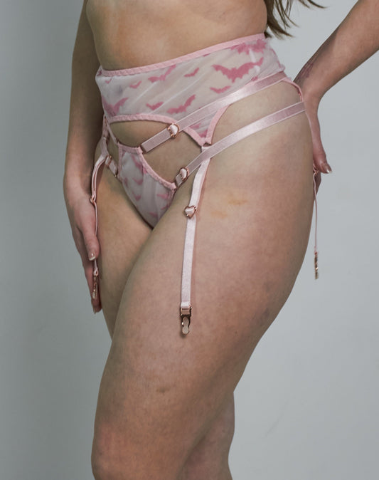 A woman modeling a light pink lingerie set with a subtle red pattern, including a bra, high-waisted briefs, and garter straps, against a plain gray studio background.
