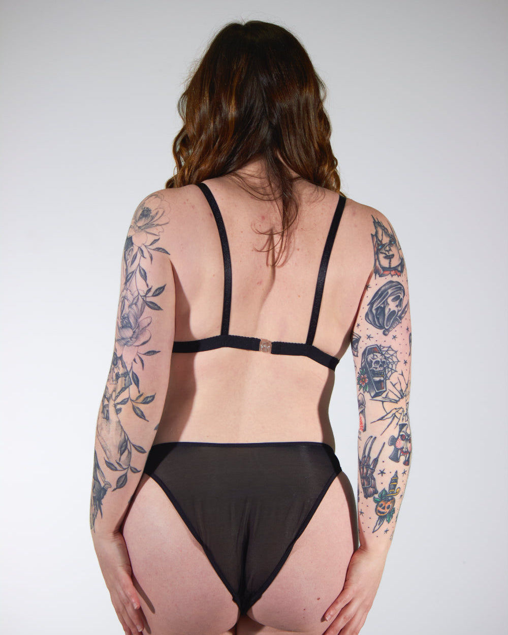 Person wearing a black strappy outfit with floral tattoos on arms and back against a white background