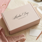Pink 'Madeline Paige intimates' box on a marble surface with lingerie and jewelry.