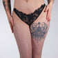 Person wearing black lace lingerie with visible tattoos on a plain background
