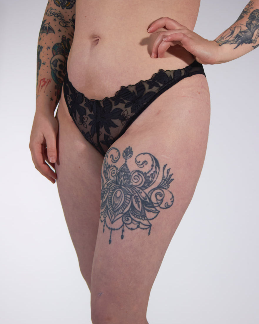 Person wearing black lace lingerie with tattoos on arms and legs against a white background